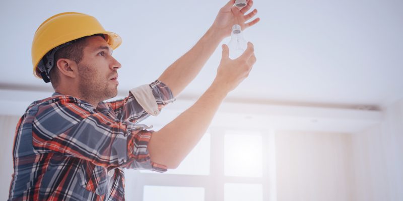 Handsome young builder in a yellow construction helmet is twisting the light bulb in. The man is looking up .